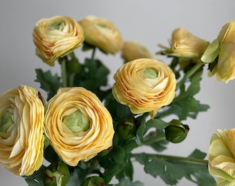May include: A bouquet of yellow ranunculus flowers with green leaves. The flowers are in full bloom and have a delicate, layered appearance.