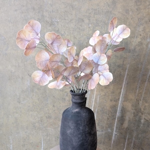 May include: A black, textured vase holds a bouquet of faux eucalyptus branches. The leaves are a mix of muted pink, beige, and light purple hues. The vase sits on a light-colored, round wooden surface against a neutral-toned background.