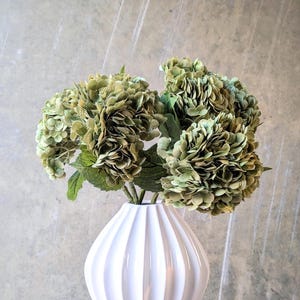 May include: A white, ribbed ceramic vase displays a bouquet of green hydrangea flowers. The vase has a rounded shape and a narrow neck. The flowers have multiple layers of petals and green leaves. The background is a neutral grey.
