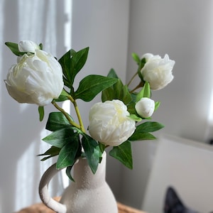 Peut inclure: Des pivoines artificielles blanches dans un vase en céramique blanche à surface texturée. Le vase a une poignée et est posé sur une surface en osier tressé.