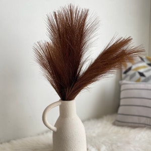 May include: A white ceramic vase with a textured surface and a handle. The vase is filled with brown dried pampas grass.