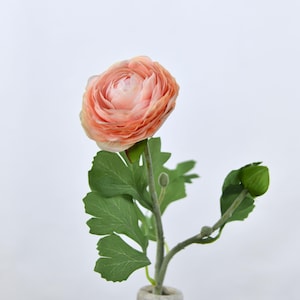 May include: A single artificial ranunculus flower in a light-coloured vase. The flower has multiple layers of peach-coloured petals. Green leaves and a bud extend from the stem. The vase is a light grey colour.