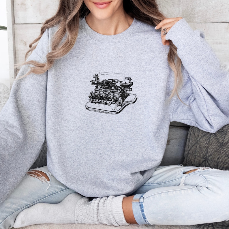 May include: A gray sweatshirt with a black and white illustration of a vintage typewriter with the text "Just Write" on the paper.