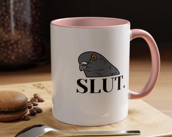 May include: White ceramic mug with a pink interior and handle. The mug features a gray pigeon graphic and the word "SLUT" in black, bold letters. A spoon and a pastry are on a wooden surface.