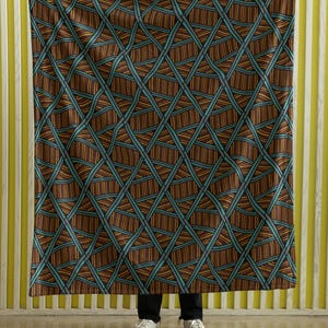 Op de afbeelding: Een patroon deken met een geometrisch ontwerp. De deken heeft een herhalend patroon van bruine, blauwgroene en oranje lijnen, waardoor een ruitvormig raster ontstaat. De deken wordt vastgehouden tegen een geel en wit gestreepte achtergrond.