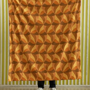 Op de afbeelding: Een deken met een retro geometrisch patroon in tinten oranje, bruin en geel. De deken wordt omhoog gehouden en toont het herhalende ontwerp van gebogen vormen. De achtergrond heeft een gestreepte muur.