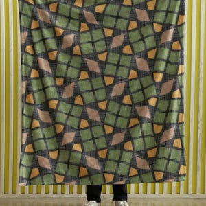 Op de afbeelding: Een grote deken met een herhalend geometrisch patroon van groene, bruine en oranje ruitvormen op een donkergrijze achtergrond. De deken wordt door een persoon omhoog gehouden, waarbij het volledige ontwerp wordt getoond. De deken is gemaakt van een zacht materiaal.