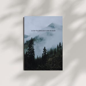 Könnte beinhalten: Eine neblige Bergszene mit einem Wald aus dunkelgrünen Bäumen. Der Text "so the lion fell in love with the lamb" steht am Himmel.
