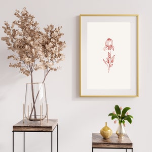 May include: A gold framed print with a red line drawing of a flower with a single stem and leaves. The print is hanging on a white wall above a brown wooden table with a clear glass vase filled with dried branches. A smaller gold vase with a bouquet of pink flowers sits on a second brown wooden table.