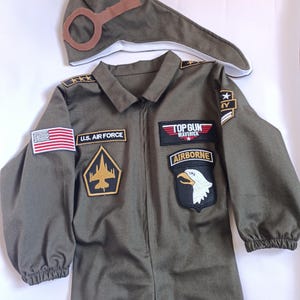 May include: Olive green Top Gun Maverick costume set. Includes a jumpsuit with patches, including a U.S. Air Force patch and an American flag, and a matching pilot's hat with goggles. The jumpsuit has a zipper closure.