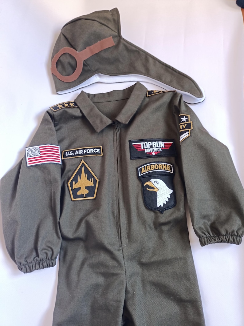 May include: Olive green Top Gun Maverick costume set. Includes a jumpsuit with patches, including a U.S. Air Force patch and an American flag, and a matching pilot's hat with goggles. The jumpsuit has a zipper closure.