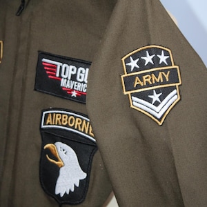 May include: Olive green flight suit with multiple patches. Patches include "TOP GUN MAVERICK" in red and white, an "AIRBORNE" patch with an eagle, and an "ARMY" patch with stars. The suit has a collar and gold star accents on the shoulder.