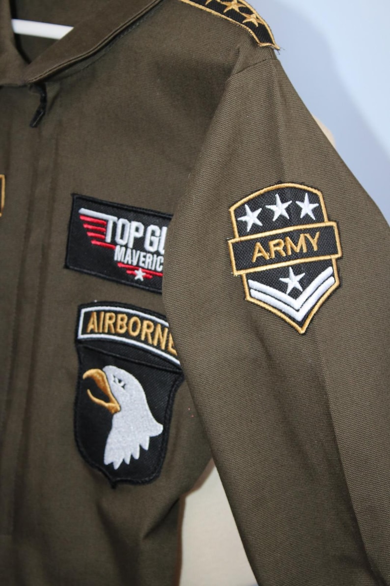 May include: Olive green flight suit with multiple patches. Patches include "TOP GUN MAVERICK" in red and white, an "AIRBORNE" patch with an eagle, and an "ARMY" patch with stars. The suit has a collar and gold star accents on the shoulder.