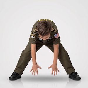 May include: A child wearing an olive green military-style uniform with patches and an American flag. The uniform includes a short-sleeved shirt and pants. The child is wearing black shoes and is bending over with their hands outstretched.