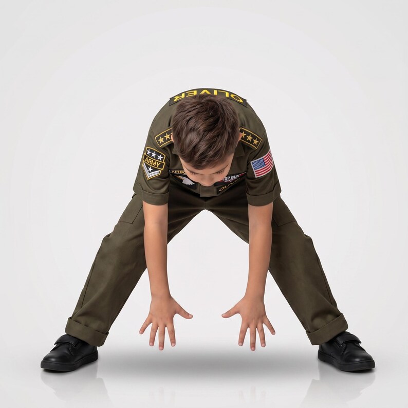 May include: A child wearing an olive green military-style uniform with patches and an American flag. The uniform includes a short-sleeved shirt and pants. The child is wearing black shoes and is bending over with their hands outstretched.