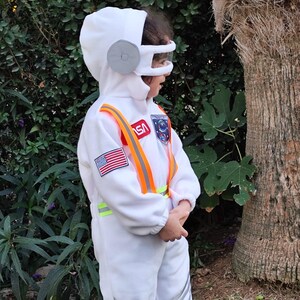 Op de afbeelding: Een kind draagt een wit astronautenkost&uuml;m met een NASA-patch, een Amerikaanse vlagpatch en oranje en gele reflecterende banden. Het kostuum heeft een witte helm met een grijs vizier.