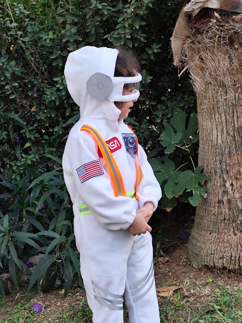 Op de afbeelding: Een kind draagt een wit astronautenkost&uuml;m met een NASA-patch, een Amerikaanse vlagpatch en oranje en gele reflecterende banden. Het kostuum heeft een witte helm met een grijs vizier.