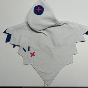 May include: A white hooded cape with blue and pink accents. The cape has a pointed bottom and jagged edges. A blue circle with a pink cross is on the hood. A pink cross and blue lines are on the cape.