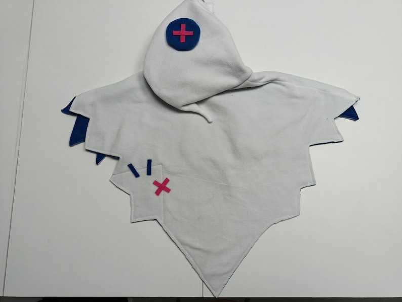 May include: A white hooded cape with blue and pink accents. The cape has a pointed bottom and jagged edges. A blue circle with a pink cross is on the hood. A pink cross and blue lines are on the cape.