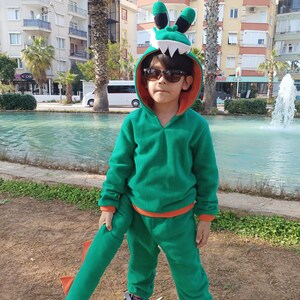Peut inclure: Un enfant portant un costume de dinosaure vert et orange avec une queue. Le costume a une capuche avec une t&ecirc;te de dinosaure et une paire de lunettes de soleil.