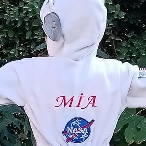 Op de afbeelding: Wit astronaut pak met een NASA-logo en de naam "MIA" geborduurd op de voorkant.
