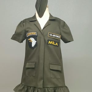 Puede incluir: Un vestido verde de estilo militar con mangas cortas y un dobladillo con volantes. Tiene parches que dicen "Airborne", "U.S. Air Force" y "Mila".