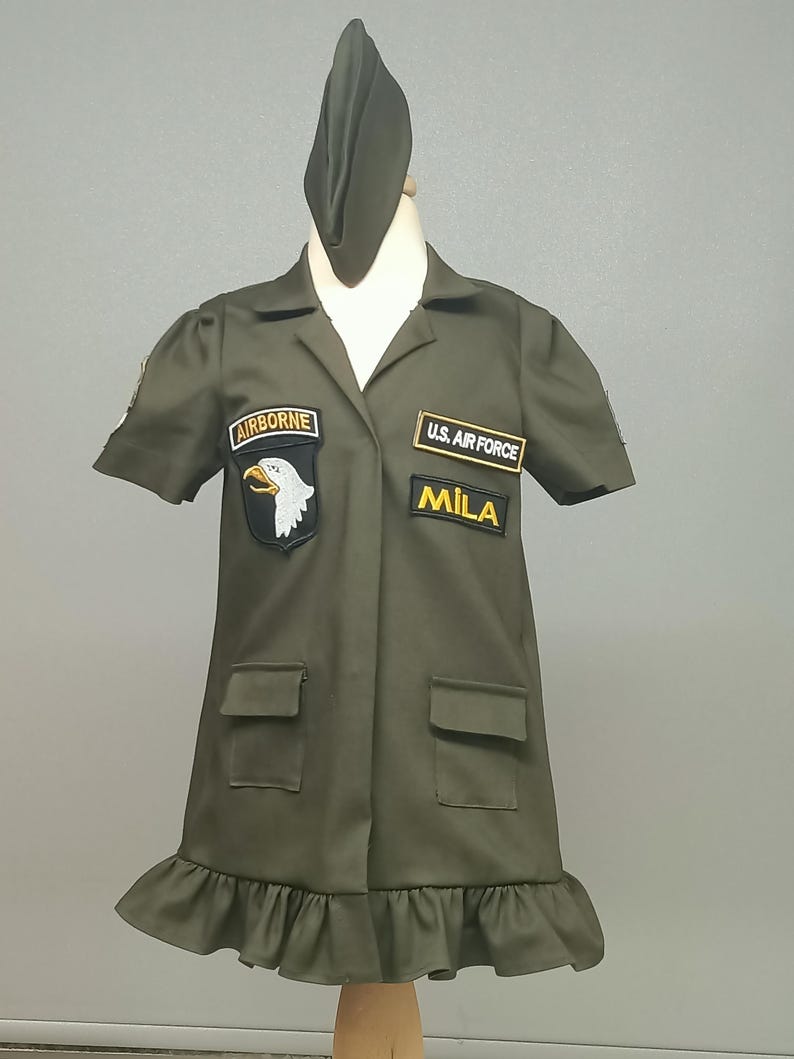 Puede incluir: Un vestido verde de estilo militar con mangas cortas y un dobladillo con volantes. Tiene parches que dicen "Airborne", "U.S. Air Force" y "Mila".