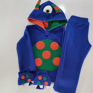 May include: A blue monster-themed fleece outfit, including a hooded sweatshirt, pants, and mittens. The sweatshirt features a large green circle with orange spots, a monster eye on the hood, and orange and green accents. The mittens have monster teeth.