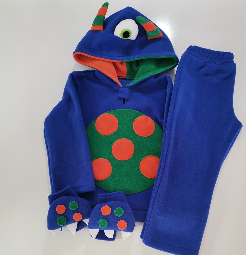 May include: A blue monster-themed fleece outfit, including a hooded sweatshirt, pants, and mittens. The sweatshirt features a large green circle with orange spots, a monster eye on the hood, and orange and green accents. The mittens have monster teeth.