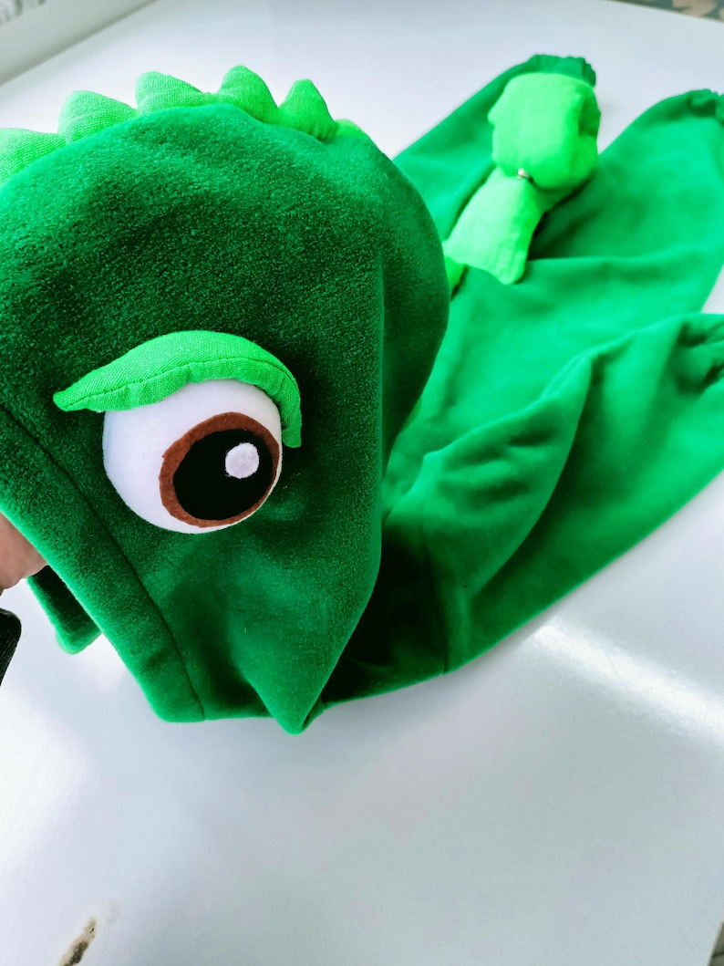 May include: Green plush dinosaur costume with a large eye and a hood. The costume is made of soft, fuzzy fabric.