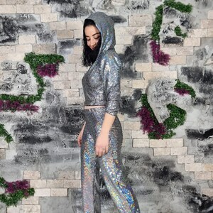 Puede incluir: Una blusa con capucha de lentejuelas plateadas y pantalones a juego. La blusa es corta y tiene una cremallera. Los pantalones son de longitud completa y tienen una pierna recta.