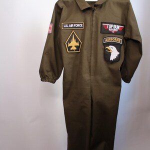 May include: Olive green jumpsuit with multiple patches, including "U.S. AIR FORCE", a gold and black emblem, and "TOP GUN MAVERICK AIRBORNE". The costume has a zipper closure and is hanging on a wooden hanger.