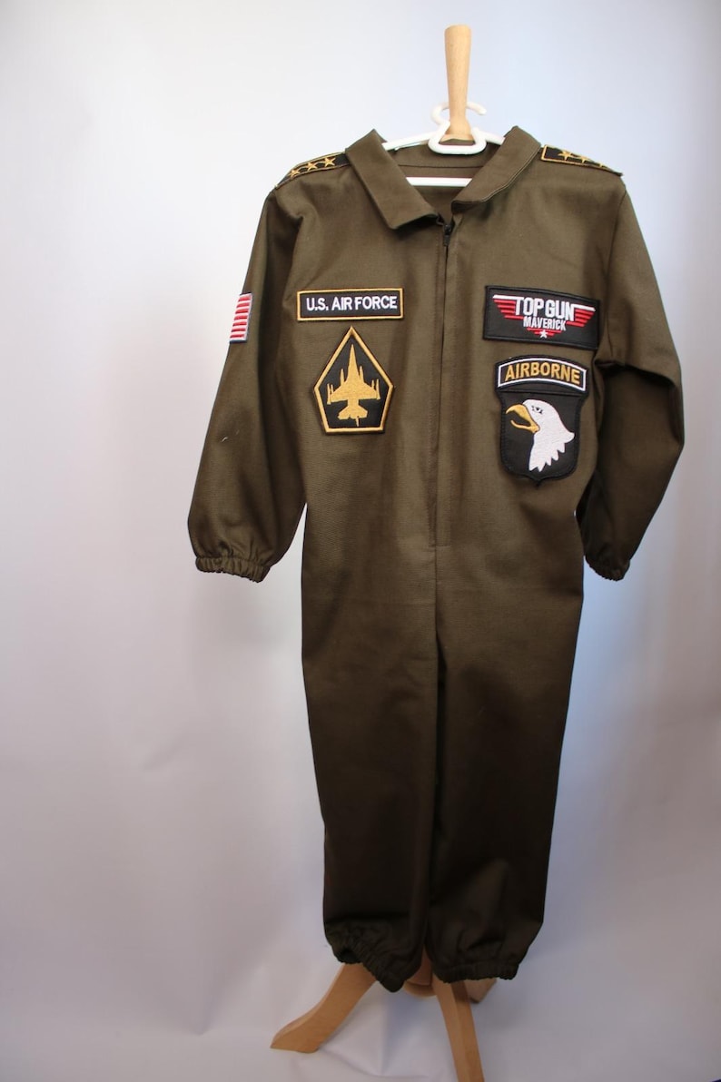 May include: Olive green jumpsuit with multiple patches, including "U.S. AIR FORCE", a gold and black emblem, and "TOP GUN MAVERICK AIRBORNE". The costume has a zipper closure and is hanging on a wooden hanger.