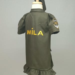 Puede incluir: Un vestido verde de estilo militar con un dobladillo con volantes y un nombre bordado en oro "MILA" en la espalda. El vestido tiene un parche en la manga derecha que dice "ARMY" con cinco estrellas.