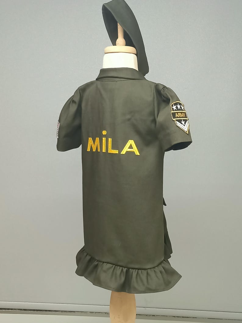 Puede incluir: Un vestido verde de estilo militar con un dobladillo con volantes y un nombre bordado en oro "MILA" en la espalda. El vestido tiene un parche en la manga derecha que dice "ARMY" con cinco estrellas.