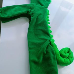May include: Green hooded chameleon costume for a child. The costume has a large green tail, a hood with a large eye, and a zipper in the front.