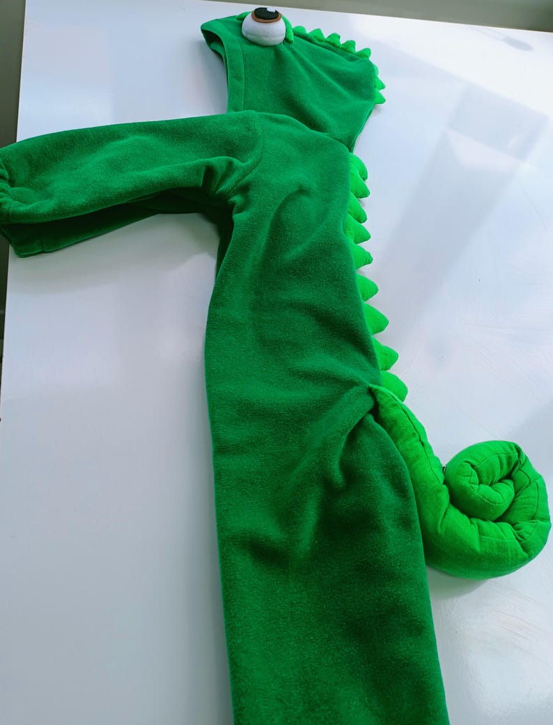 May include: Green hooded chameleon costume for a child. The costume has a large green tail, a hood with a large eye, and a zipper in the front.