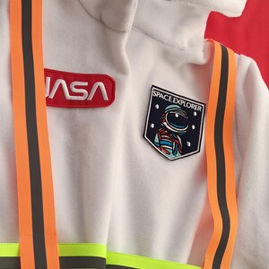 Op de afbeelding: Wit astronaut kostuum met oranje en zwarte banden, een NASA-patch en een Space Explorer-patch met een astronaut ontwerp.