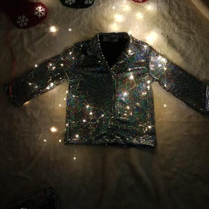 May include: A silver sequined jacket with long sleeves and a collar, illuminated by fairy lights. The jacket is the focal point, set against a soft, textured background. Christmas stockings with snowflake designs are in the background.