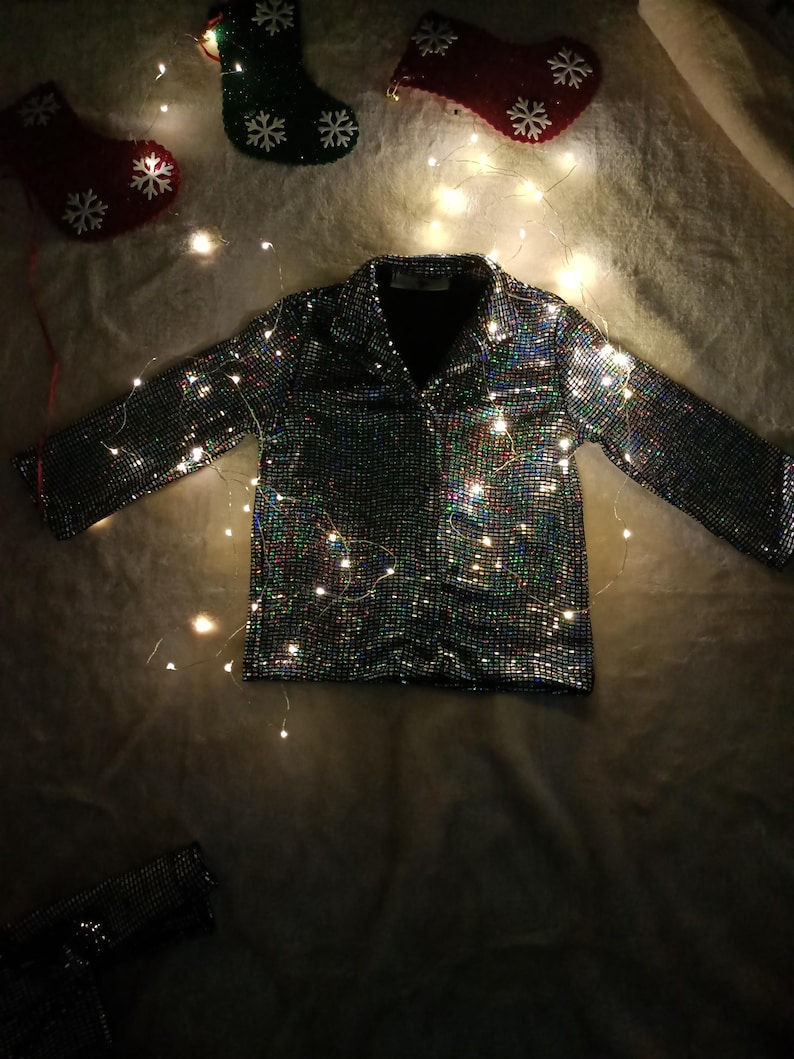 May include: A silver sequined jacket with long sleeves and a collar, illuminated by fairy lights. The jacket is the focal point, set against a soft, textured background. Christmas stockings with snowflake designs are in the background.
