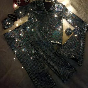 May include: A matching pajama set with a jacket, pants, eye mask, and scrunchie. The set is covered in iridescent sequins, reflecting multiple colors. The pajama top has a collared neckline and button closure. Christmas stockings and fairy lights are in the background.