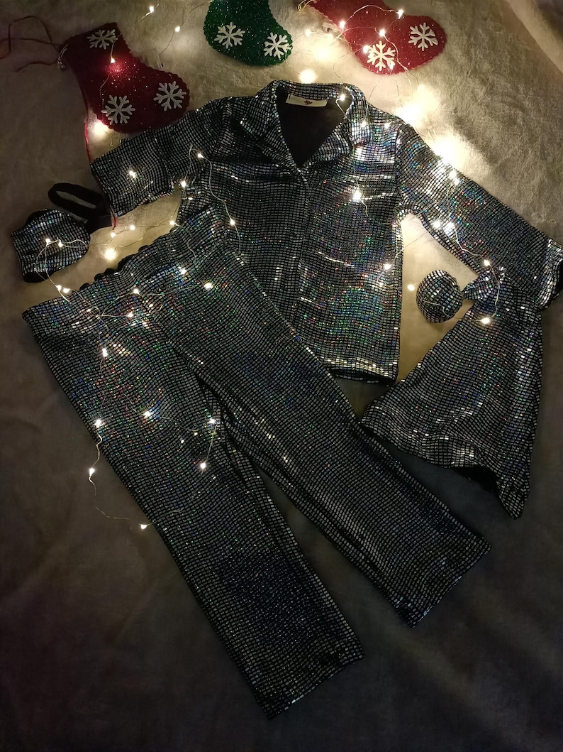May include: A matching pajama set with a jacket, pants, eye mask, and scrunchie. The set is covered in iridescent sequins, reflecting multiple colors. The pajama top has a collared neckline and button closure. Christmas stockings and fairy lights are in the background.
