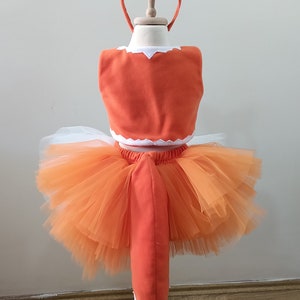 May include: Orange and white fox costume for a child. The costume includes a tutu skirt, a top with a tail, and a headband with fox ears.