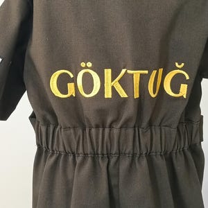 Puede incluir: Mono verde oliva oscuro con la palabra "G&Ouml;KTUĞ" bordada en hilo dorado en la espalda. El mono tiene cintura el&aacute;stica y mangas cortas. La tela parece ser un material duradero, de estilo trabajo.