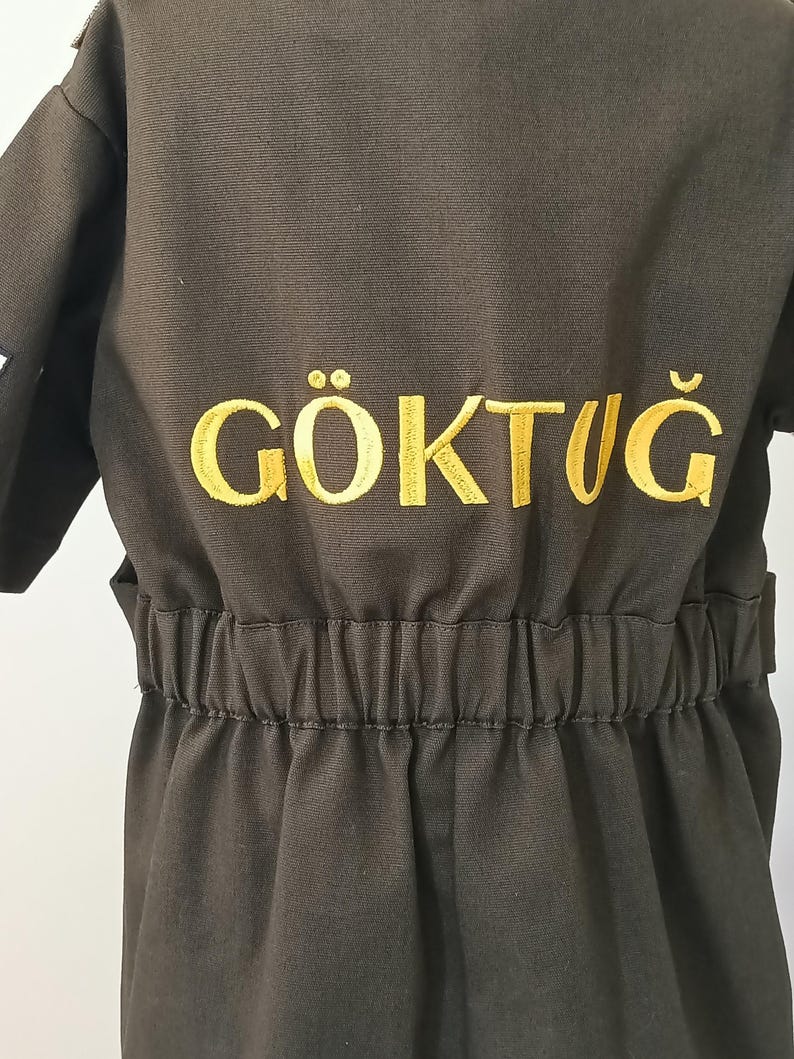 Puede incluir: Mono verde oliva oscuro con la palabra "G&Ouml;KTUĞ" bordada en hilo dorado en la espalda. El mono tiene cintura el&aacute;stica y mangas cortas. La tela parece ser un material duradero, de estilo trabajo.
