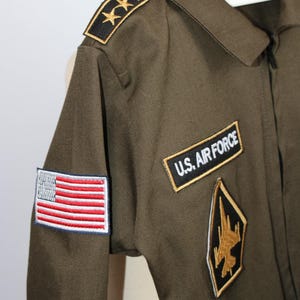 May include: Olive green U.S. Air Force flight suit with embroidered patches. Features an American flag patch, "U.S. AIR FORCE" patch, and a gold and black emblem. Includes gold star epaulettes.