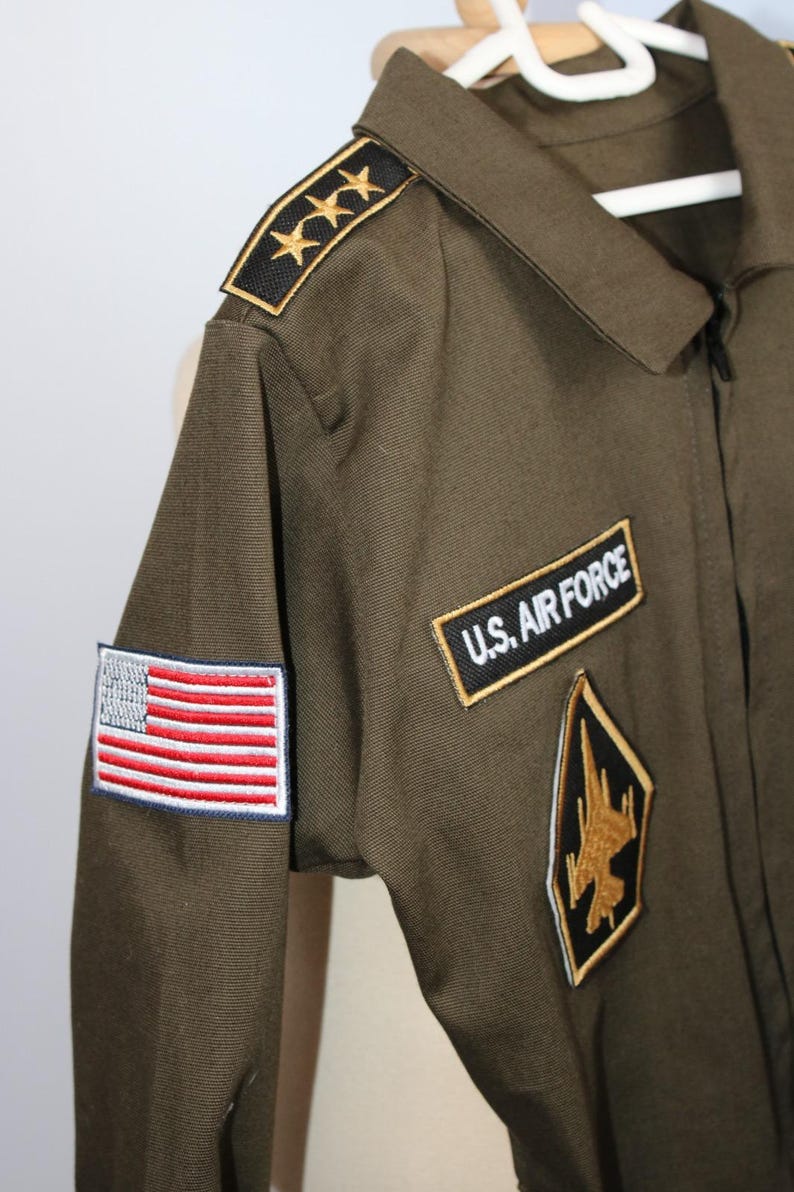 May include: Olive green U.S. Air Force flight suit with embroidered patches. Features an American flag patch, "U.S. AIR FORCE" patch, and a gold and black emblem. Includes gold star epaulettes.