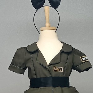 May include: Olive green costume dress with short sleeves, a black ribbon belt, and gold buttons. The dress has a collar and a name tag that reads "Mary". A black satin bow is attached to a headband.