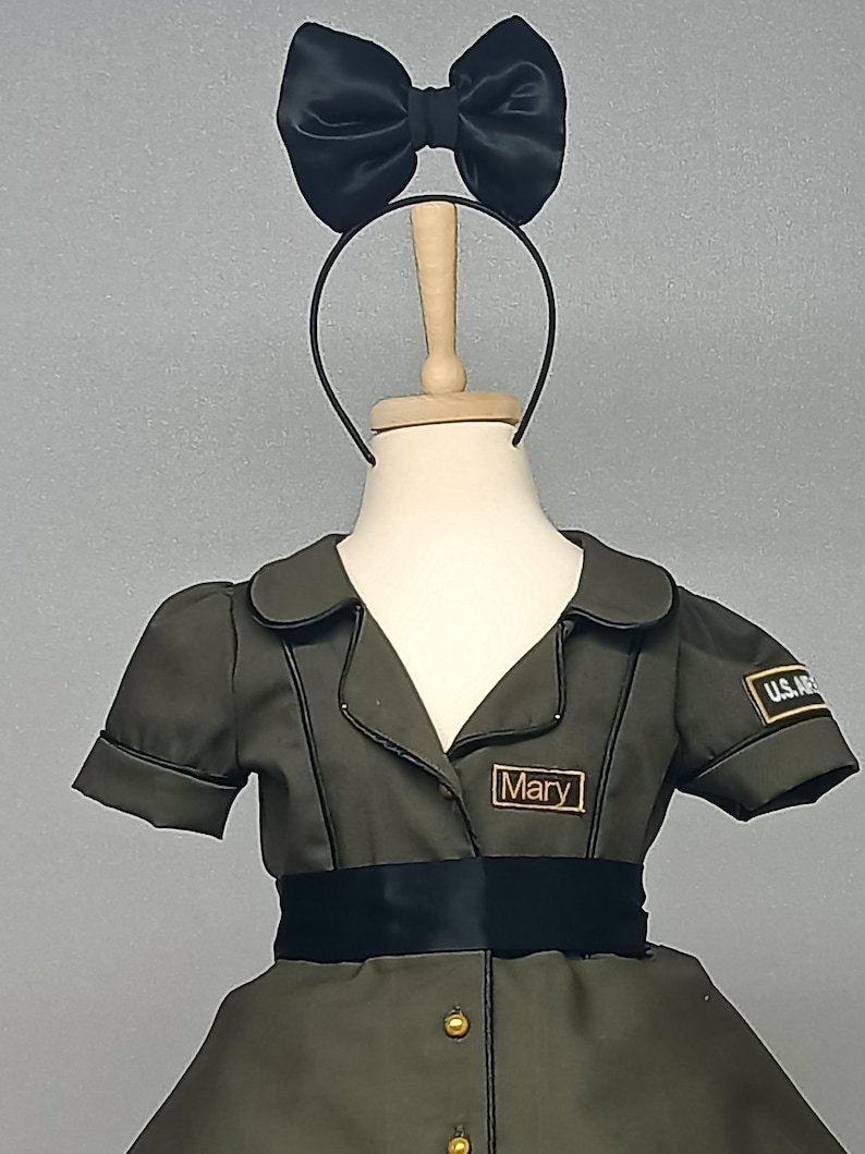 May include: Olive green costume dress with short sleeves, a black ribbon belt, and gold buttons. The dress has a collar and a name tag that reads "Mary". A black satin bow is attached to a headband.