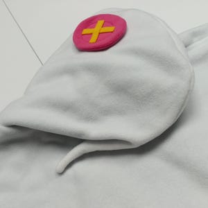 May include: White fleece hat with a pink cross emblem. The hat has a rounded shape with a pointed tail. The cross emblem is a pink circle with a yellow cross in the center. The hat is made of a soft, plush material.